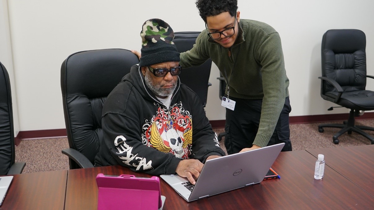 Jordan Owens, Computer Program Assistant, stands with an HACP resident who is typing on an open laptop.
