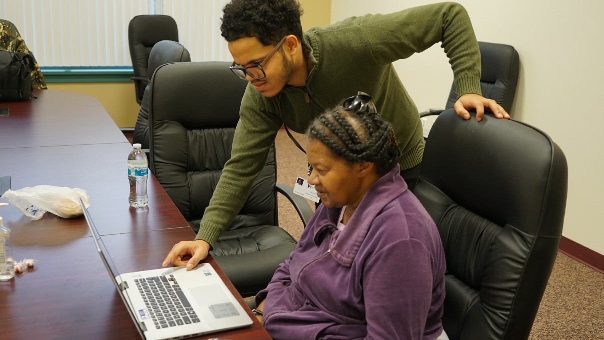 Jordan Owens, Computer Program Assistant, stands with an HACP resident who is sitting in front of an open laptop.