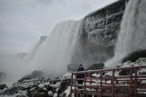 Christine stands smiling, in winter coat and accessories, on a platform in front of the niagra falls on a cloudy, grey day