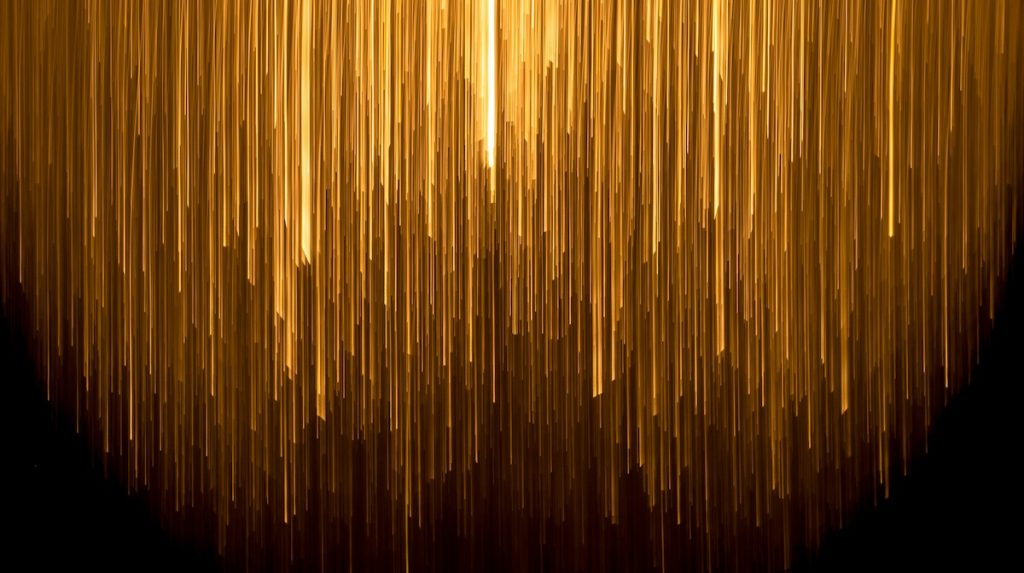 Thin gold columns of light of different shades and lengths against a dark background.