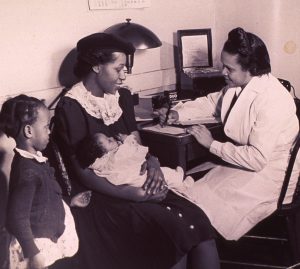 An African American nurse is recording a medical history as it is reported by a woman holding an infant.