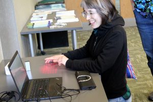 Maddie (author of this post) kneeling at a table, attempting to use a projected keyboard, and Smyle Mouse software on a laptop