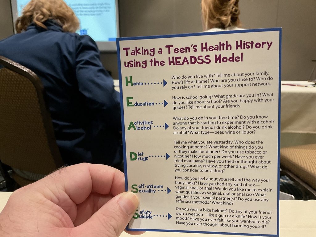 A hand holding a flyer titled "Taking a Teen's Health History using the HEADSS Model" along with step-by-step directions.