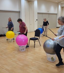 Four women participating in a cardio drumming class using drumsticks and baskets to hold inflatable balls. 