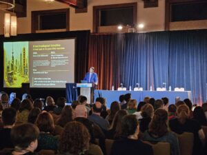 Speaker standing at a podium with a large screen to their right and attendees seated at tables in the foreground.