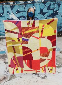 Maddie holding up a colorful quilt of improvisational curves in warm tones. Background is a blue graffitied wall.