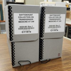 Two gray archival boxes sit side by side on a brown desk. The boxes are labeled "Dayton/Hunt Collection on Transgender Medicine at UW."