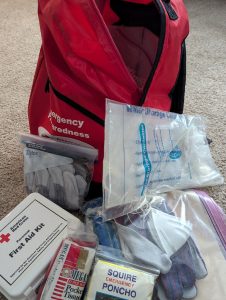 Red emergency preparedness backpack with some contents on display including a first aid kit, poncho, gloves, tissues, etc.