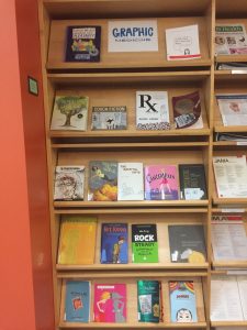 Picture of the graphic medicine collection at Dana Library at UVM (front facing book shelves with about 20 graphic medicine book covers visible including books such as Are you My Mohter, Rx, Cancer Vixen, Hey, Kiddo and more)