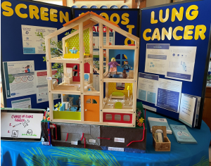 Health fair exhibit set-up on a blue table cloth with a board in teh background with information about preventing and screening for lung cancer. In the foreground is a wooden doll house set-up to demonstrate information about where radon occurs in homes and the tesing and abatement process.