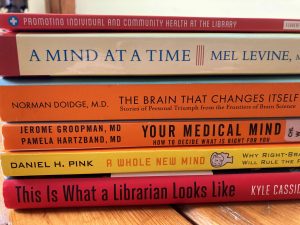 Stack of library books with the titles Promoting individual and community health at the library, a mind at a time, The brain that changes itself, your medical mind, a whole new mind, This is what a librarian looks like.