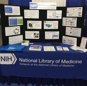 NNLM exhibit materials for APHA 2022 with a blue table cloth reading "National Library of Medicine, Network of the National Library of Medicine" handouts on the table and a display board with information about NNLM classes for Public Health audiences, NNLM overview and a PHDL overview.