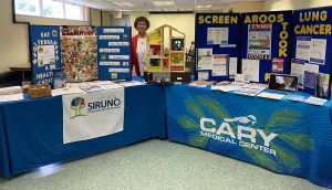 Nancy Holmquist standing between two tables with health information displayed. On the left hand table is information about stroke prevention and the Mediterranean Diet, in the middle is a dollhouse with information about Radon and on the table on the right is information about lung cancer including screening, prevention and radon as a cause of lung cancer.