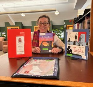 Tatjana Saccio standing behind a bookshelf holding up a copy of A Fade of Light with Tangles on the right, a binder of health information to the left and a media bag in front.