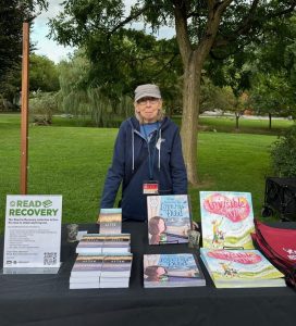 Mary Ratzer behind a table with resources from Read to Recovery. In the background is a green field and trees.